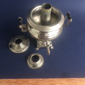 Puede incluir: Un samovar plateado con cuerpo redondo, chimenea central y grifo. Tiene dos asas con detalles negros y se asienta sobre una peque&ntilde;a base. Dos piezas separadas est&aacute;n al lado sobre una superficie azul.