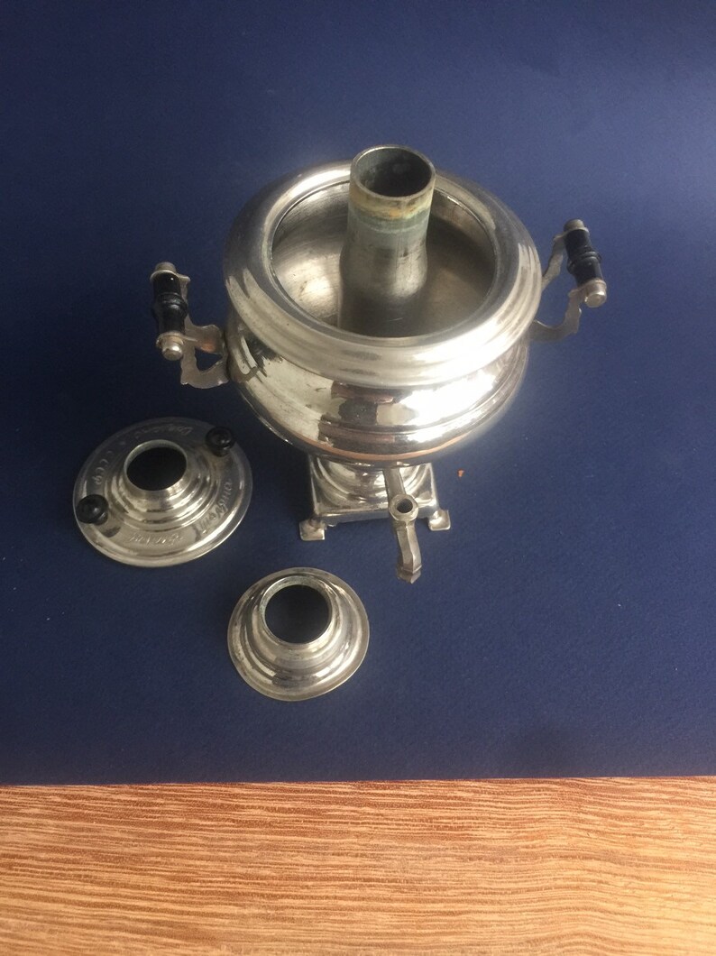Puede incluir: Un samovar plateado con cuerpo redondo, chimenea central y grifo. Tiene dos asas con detalles negros y se asienta sobre una peque&ntilde;a base. Dos piezas separadas est&aacute;n al lado sobre una superficie azul.