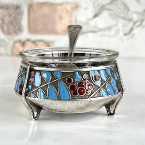 May include: A silver salt cellar with a glass top and three legs. The cellar is decorated with a blue and red floral pattern.