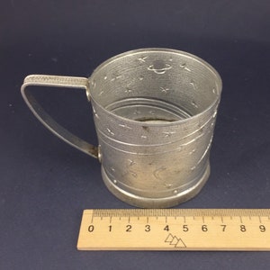 Peut inclure: Une tasse en m&eacute;tal argent&eacute; avec une poign&eacute;e et un motif sur le th&egrave;me de l'espace. La tasse est d&eacute;cor&eacute;e d'&eacute;toiles, d'un croissant de lune et d'une plan&egrave;te. La tasse est pos&eacute;e sur une r&egrave;gle en bois.