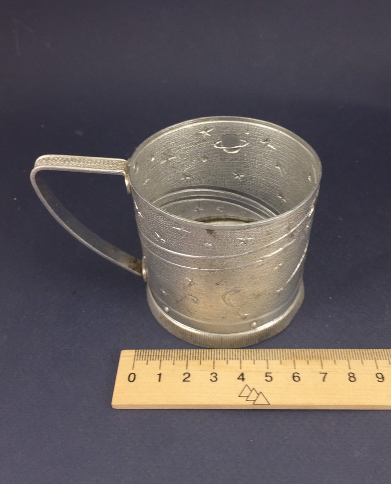 Peut inclure: Une tasse en m&eacute;tal argent&eacute; avec une poign&eacute;e et un motif sur le th&egrave;me de l'espace. La tasse est d&eacute;cor&eacute;e d'&eacute;toiles, d'un croissant de lune et d'une plan&egrave;te. La tasse est pos&eacute;e sur une r&egrave;gle en bois.