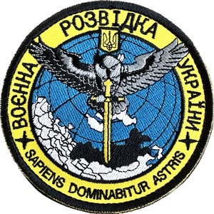 Puede incluir: Un parche bordado amarillo y negro con un globo terráqueo azul, un águila plateada con una espada dorada y el texto "РОЗВІДКА УКРАЇНИ" en cirílico ucraniano. El texto "SAPIENS DOMINABITUR ASTRIS" también es visible en la parte inferior del parche.