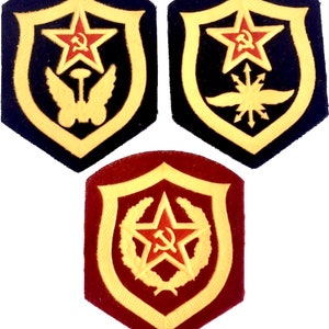 Puede incluir: Tres parches bordados con una estrella roja y un emblema de martillo y hoz. Los parches tienen forma de escudo con un borde dorado. El primer parche tiene un fondo negro con un cohete alado dorado. El segundo parche tiene un fondo negro con una estrella alada dorada. El tercer parche tiene un fondo marrón con una corona de laurel dorada.