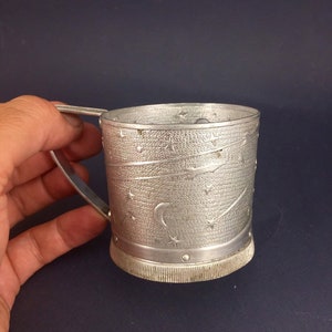 Peut inclure: Une tasse en m&eacute;tal argent&eacute; avec une poign&eacute;e et un motif d'&eacute;toiles et de croissant de lune. La tasse a une surface textur&eacute;e et un bord l&eacute;g&egrave;rement sur&eacute;lev&eacute;.