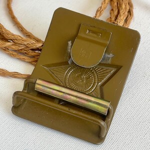 May include: An olive-green metal military belt buckle with a star emblem and a rope cord. The buckle has a rectangular shape with a hinged clasp and a metal bar. The star emblem is in the center of the buckle.