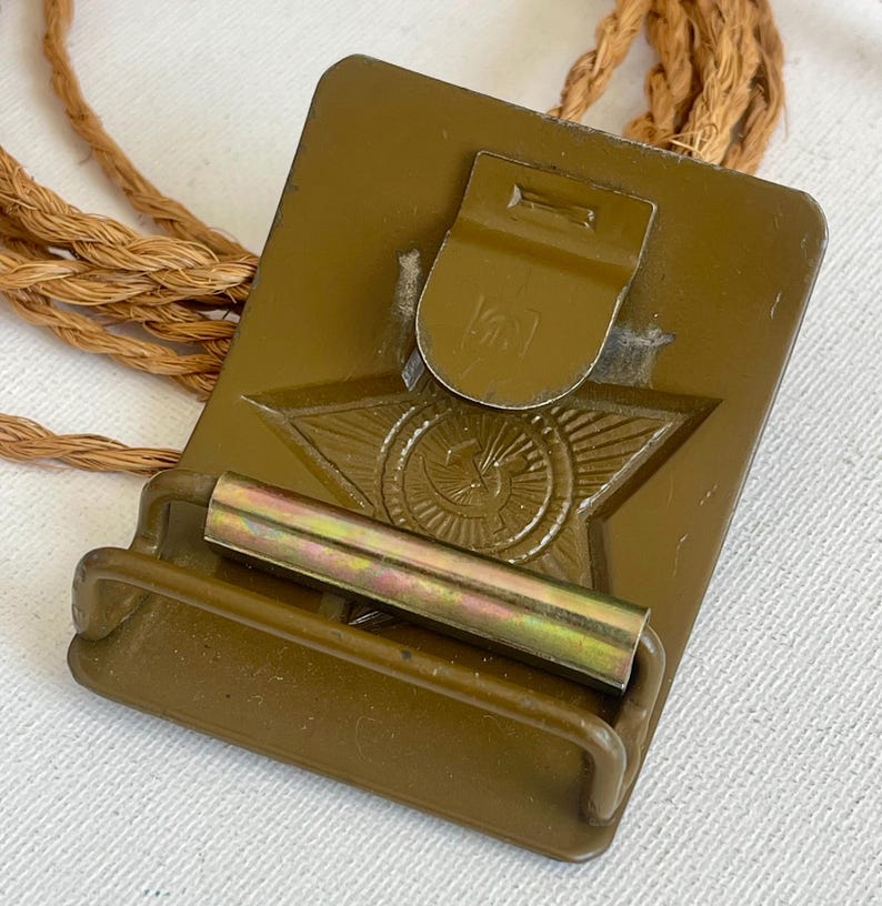 May include: An olive-green metal military belt buckle with a star emblem and a rope cord. The buckle has a rectangular shape with a hinged clasp and a metal bar. The star emblem is in the center of the buckle.