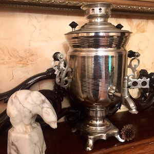 May include: A silver samovar with ornate handles and a spout, sitting on a wooden surface with a white marble bear figurine.