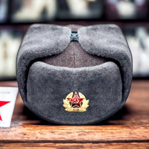 Może przedstawiać: Szary futrzaną czapka uszanka z czerwoną gwiazdą i złotym emblematem z przodu.