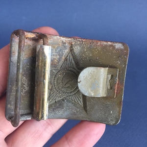 Pu&ograve; includere: Una fibbia per cintura in metallo verde con un design a stella, martello e falce. La fibbia &egrave; usurata e ha uno scorrevole in ottone.