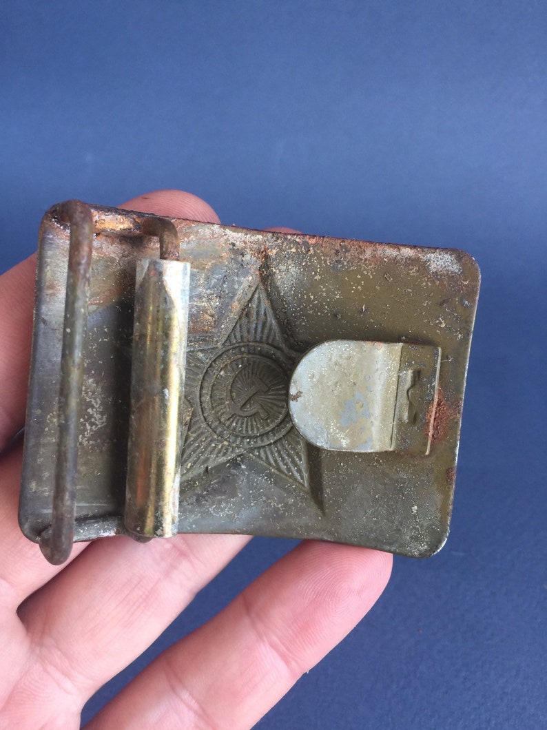 Pu&ograve; includere: Una fibbia per cintura in metallo verde con un design a stella, martello e falce. La fibbia &egrave; usurata e ha uno scorrevole in ottone.