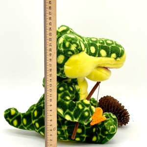 May include: A green and yellow plush alligator toy holding a fishing rod and a small orange fish. The alligator is sitting on a white surface with a pine cone next to it.
