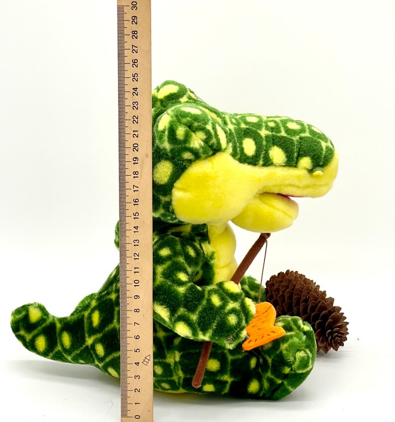 May include: A green and yellow plush alligator toy holding a fishing rod and a small orange fish. The alligator is sitting on a white surface with a pine cone next to it.