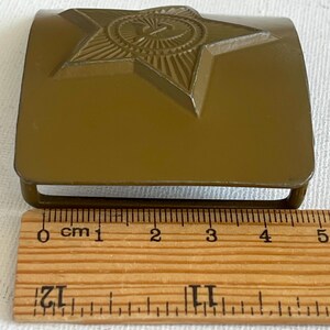 May include: Olive green metal belt buckle featuring a raised star emblem with a hammer and sickle. The buckle measures approximately 2 inches in length and is a vintage military accessory.