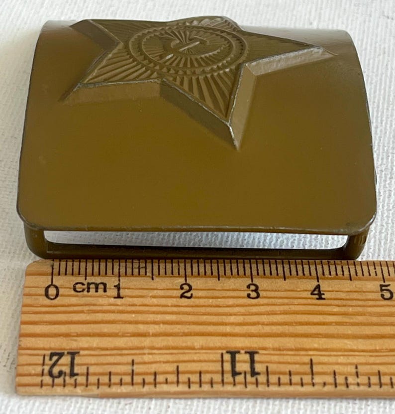 May include: Olive green metal belt buckle featuring a raised star emblem with a hammer and sickle. The buckle measures approximately 2 inches in length and is a vintage military accessory.