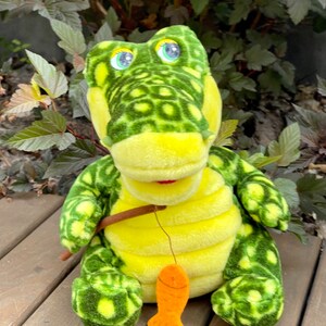 May include: A green and yellow plush crocodile toy holding a fishing rod with an orange fish attached. The crocodile has large, round eyes and a toothy grin.
