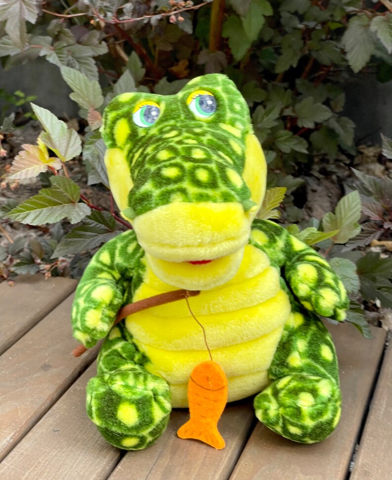 May include: A green and yellow plush crocodile toy holding a fishing rod with an orange fish attached. The crocodile has large, round eyes and a toothy grin.