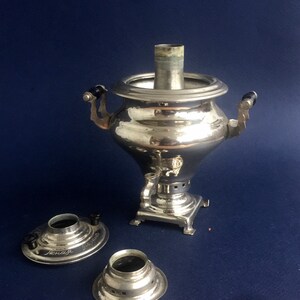 Puede incluir: Un samovar plateado brillante con asas negras y un pico, sobre un fondo azul oscuro. Dos piezas plateadas separadas con perillas negras y texto est&aacute;n delante. Un aparato tradicional para calentar agua o t&eacute;.