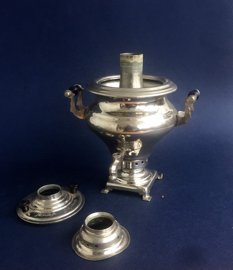 Puede incluir: Un samovar plateado brillante con asas negras y un pico, sobre un fondo azul oscuro. Dos piezas plateadas separadas con perillas negras y texto est&aacute;n delante. Un aparato tradicional para calentar agua o t&eacute;.