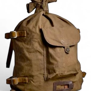 May include: A brown canvas backpack with leather straps and a large flap pocket. The backpack has a drawstring closure at the top and a buckle on the front flap.