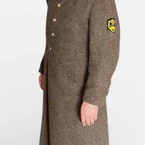 Puede incluir: Un abrigo de lana marrón con botones dorados e insignias en los hombros. El abrigo tiene un diseño cruzado y un cuello negro. Un parche amarillo y negro es visible en la manga superior izquierda. Estilo militar vintage.