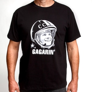 Puede incluir: Camiseta negra con un gráfico blanco de Yuri Gagarin, el primer humano en el espacio, usando un casco con las letras "CCCP" en él. El texto "GAGARIN" está impreso debajo de la imagen.