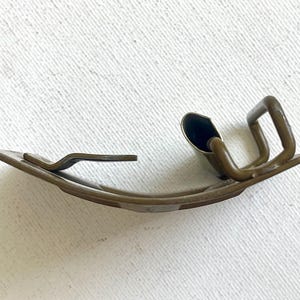 May include: A curved, antique-looking metal clip with a dark bronze finish. The clip has a rectangular base with a curved design and a raised, looped section. The clip is designed to hold something in place.
