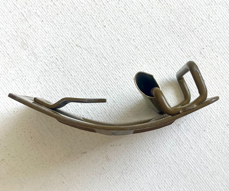May include: A curved, antique-looking metal clip with a dark bronze finish. The clip has a rectangular base with a curved design and a raised, looped section. The clip is designed to hold something in place.