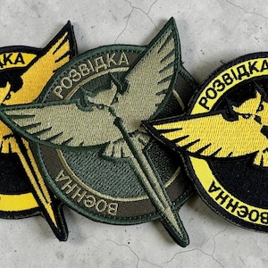 May include: Embroidered patches featuring a central design of a stylized bird with a sword, surrounded by text. The patches are primarily olive green, black, and yellow, with the text in white. The patches are likely military or tactical in nature.