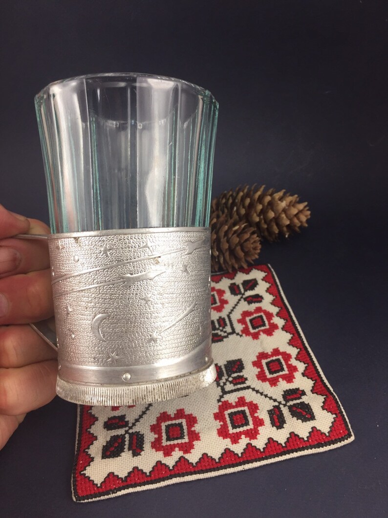 Peut inclure: Un porte-verre en argent avec un motif d&eacute;coratif d'&eacute;toiles et d'un croissant de lune. Le porte-verre est en m&eacute;tal et a une surface textur&eacute;e. Le porte-verre est pos&eacute; sur un tissu brod&eacute; rouge et blanc.