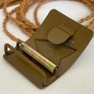 May include: A vintage olive drab metal belt buckle with a star emblem. The buckle has a rectangular shape with a hinged clasp and a roller bar. A length of brown rope is in the background.