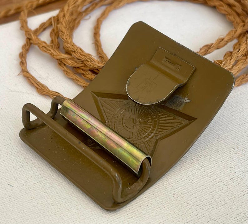 May include: A vintage olive drab metal belt buckle with a star emblem. The buckle has a rectangular shape with a hinged clasp and a roller bar. A length of brown rope is in the background.