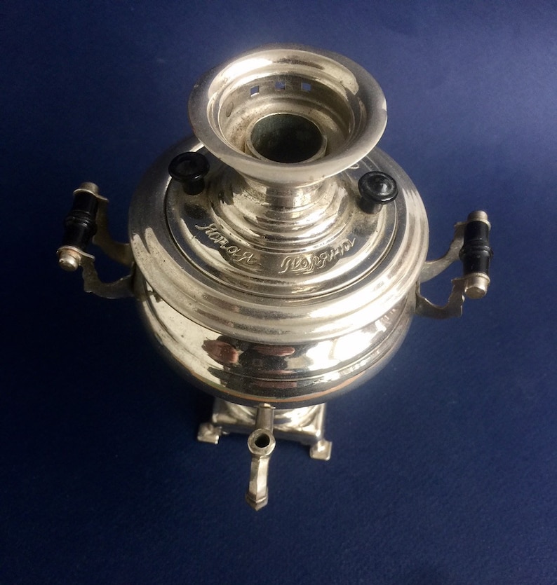 Puede incluir: Un samovar plateado con asas negras y un pico. El samovar tiene un cuerpo redondo y una parte superior acampanada. El texto "Новая Поляна" est&aacute; grabado en la tapa. El samovar est&aacute; sobre una superficie azul oscuro.