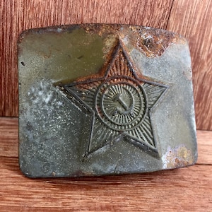 Pu&ograve; includere: Una fibbia per cintura in metallo vintage con una finitura verde usurata. La fibbia presenta una grande stella rossa con un martello e una falce all'interno di un cerchio al centro.