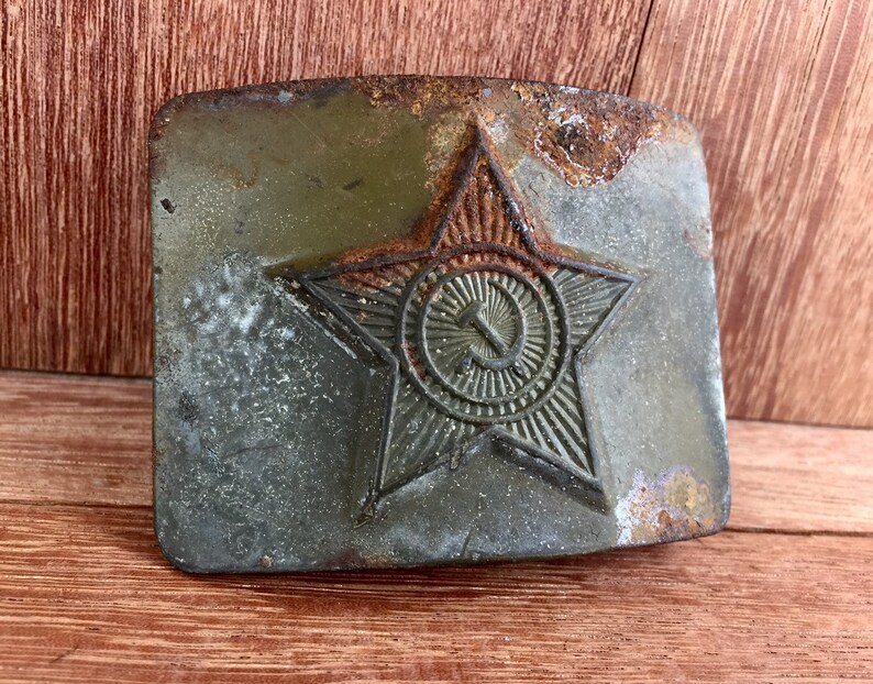 Pu&ograve; includere: Una fibbia per cintura in metallo vintage con una finitura verde usurata. La fibbia presenta una grande stella rossa con un martello e una falce all'interno di un cerchio al centro.