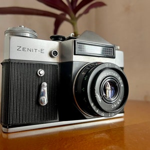 Puede incluir: Una cámara de película Zenit-E plateada y negra con un objetivo Industar-50-2 negro. La cámara tiene una cubierta de imitación de cuero negro y una placa superior plateada con un fotómetro incorporado.