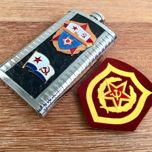 May include: A silver flask with a black leather-like exterior featuring three red star emblems. The flask is on a wooden surface next to a red and gold embroidered patch with a red star and hammer and sickle.