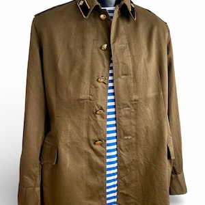 Könnte beinhalten: Eine olivgrüne Jacke im Militärstil mit goldfarbenen Knöpfen und einem schwarzen Kragen mit goldenen Akzenten. Die Jacke ist zugeknöpft und darunter ist ein blau-weiß gestreiftes Hemd zu sehen. Die Jacke hat zwei Vordertaschen und lange Ärmel.