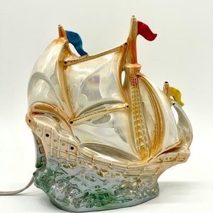 Puede incluir: Una lámpara de cerámica con forma de barco con un esmalte blanco iridiscente. El barco tiene un casco marrón, una vela blanca y tres banderas de colores. La lámpara está sobre una base verde y blanca.
