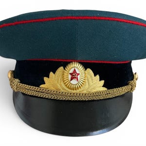 May include: A dark green military hat with a red stripe along the top edge. The hat features a black brim, gold trim, and a gold emblem with a red star. This vintage hat is likely of historical significance.
