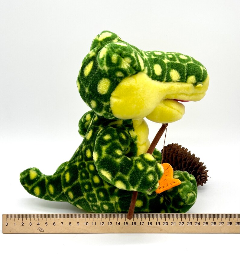 May include: A green and yellow plush alligator toy holding a fishing rod and a fish. The alligator has a spotted pattern and is sitting on a white background.