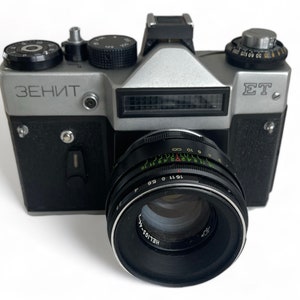 Puede incluir: Una cámara Zenit ET plateada y negra con un objetivo Helios-44. La cámara tiene una cubierta de imitación de cuero negro y una placa superior plateada con el nombre de la marca "Zenit" y el nombre del modelo "ET" impreso en ella.