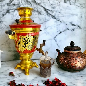 Könnte beinhalten: Ein gold-roter Samowar mit schwarzen Akzenten, eine dekorative Teekanne und ein Glas Tee in einem Metallhalter. Der Samowar hat florale Muster. Die Teekanne ist braun mit goldenen Akzenten.
