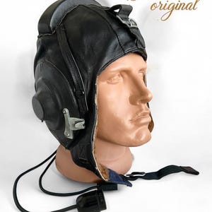 May include: A black leather aviator helmet with earmuffs and a microphone. The helmet has a brown fur lining and a chin strap. The text "Guaranteed 100% original" and "Made in USSR, 1974." is visible in the image.
