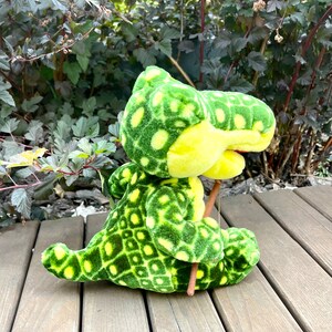 May include: Green and yellow plush crocodile toy holding a brown stick. The crocodile has a patterned design on its body.