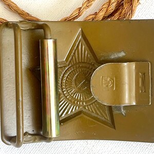 May include: A vintage olive-drab metal belt buckle featuring a raised star emblem with a central circular design. The buckle has a rectangular shape with a hinged bar and a metal loop. A brown rope is visible.
