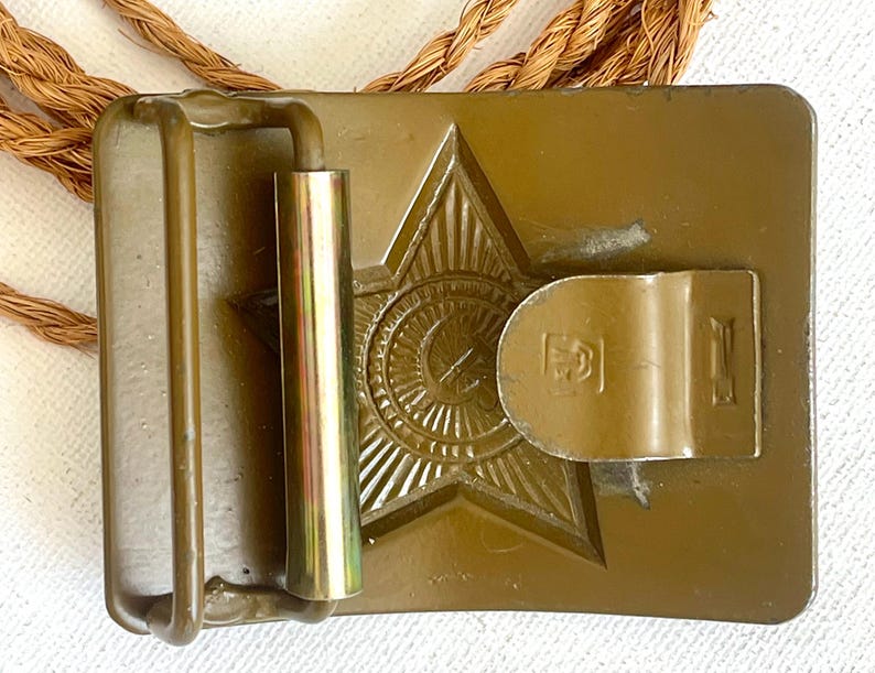 May include: A vintage olive-drab metal belt buckle featuring a raised star emblem with a central circular design. The buckle has a rectangular shape with a hinged bar and a metal loop. A brown rope is visible.