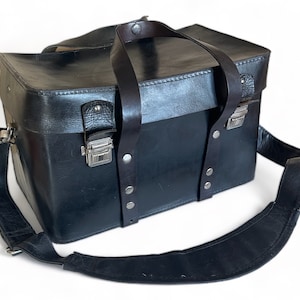 May include: A black leather camera bag with a shoulder strap and two top handles. The bag has metal clasps and rivets.