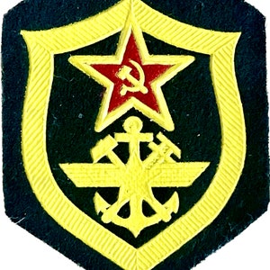 May include: A yellow and black embroidered patch with a red star and a hammer and sickle inside the star. The star is above a yellow anchor with wings.
