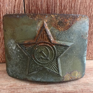 Pu&ograve; includere: Una fibbia per cintura in metallo verde ruggine con una stella rossa e una falce e martello all'interno della stella.