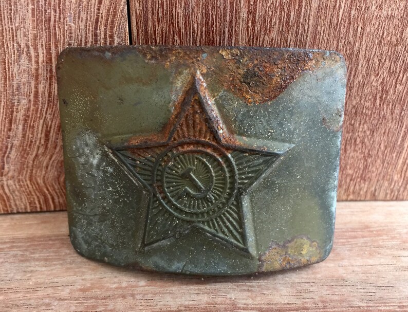 Pu&ograve; includere: Una fibbia per cintura in metallo verde ruggine con una stella rossa e una falce e martello all'interno della stella.